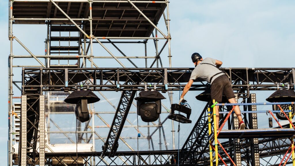 Installation of lighting equipment on the concert stage. A worker installs a powerful lantern on a metal frame under the open sky. Preparation for mass events. Unrecognizable person